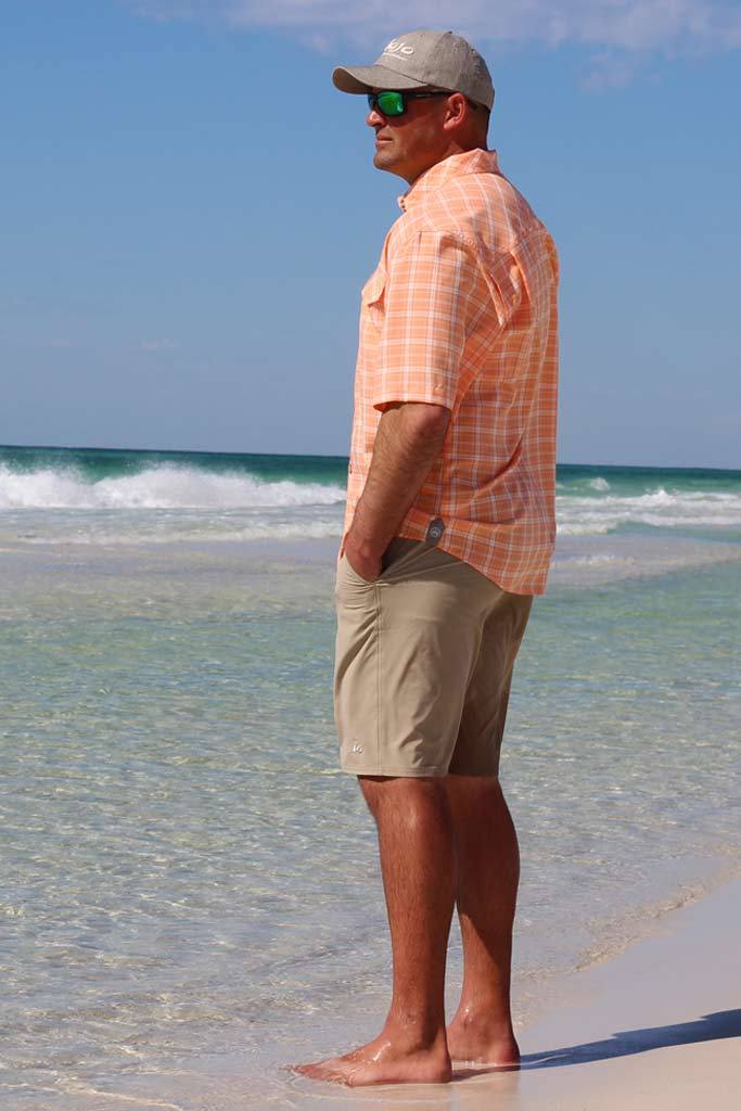Man standing on a beach wearing an orange checkered shirt and beige shorts.