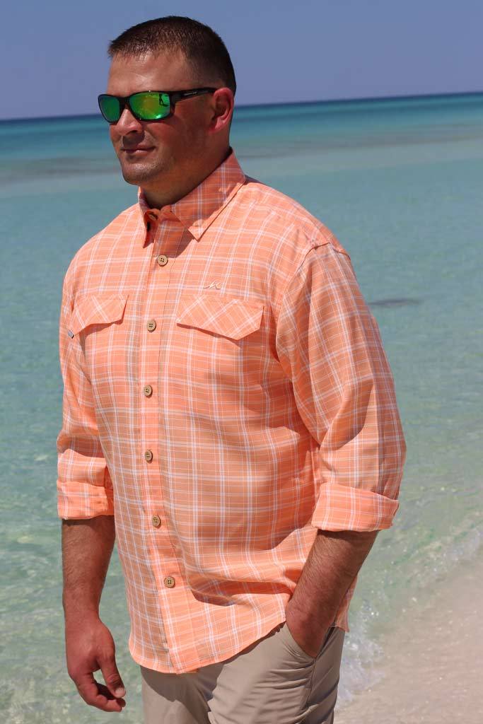 Man wearing an orange plaid shirt standing on a beach with clear blue water.