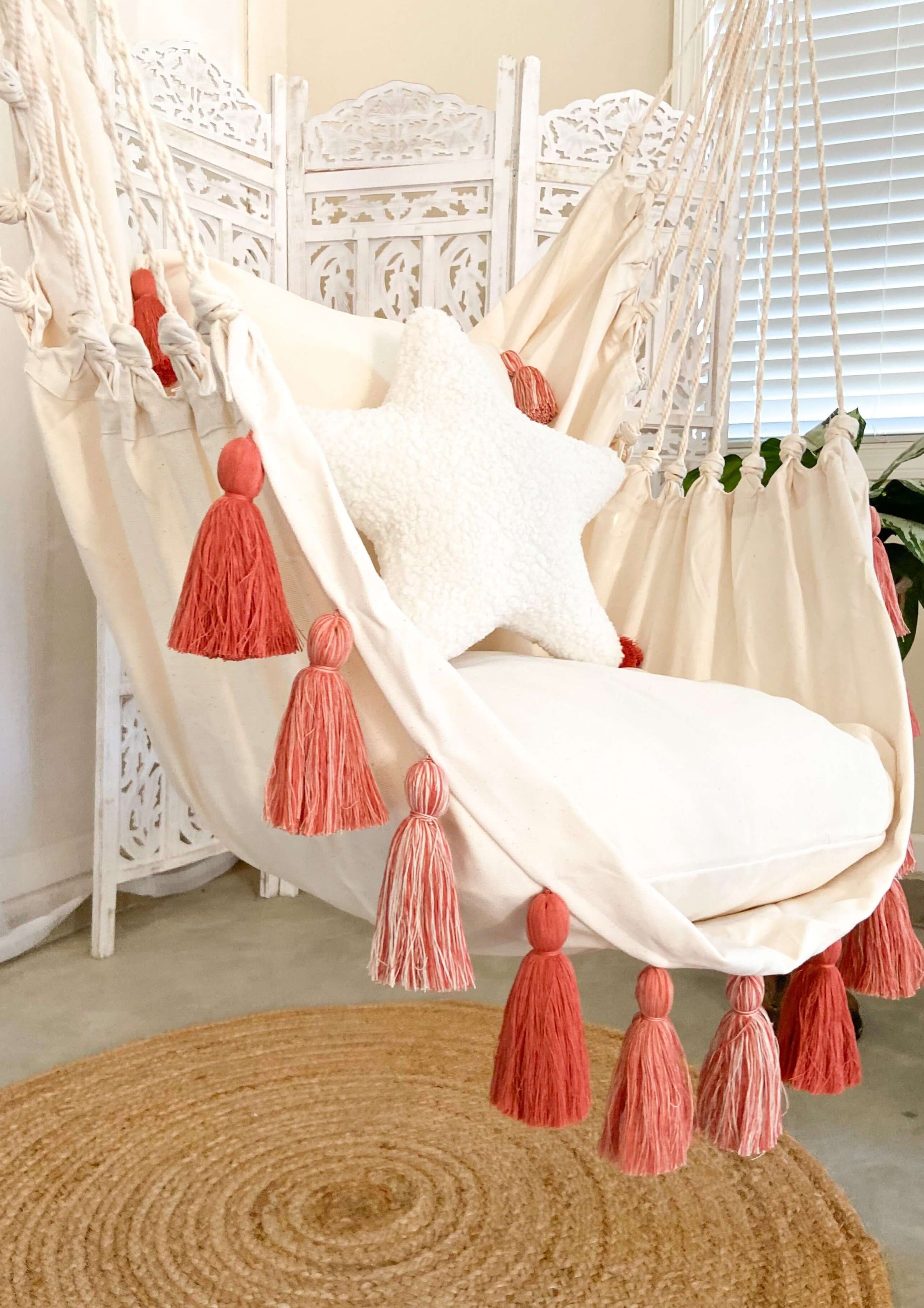 White hammock chair with coral tassels in a room with a rug and window.