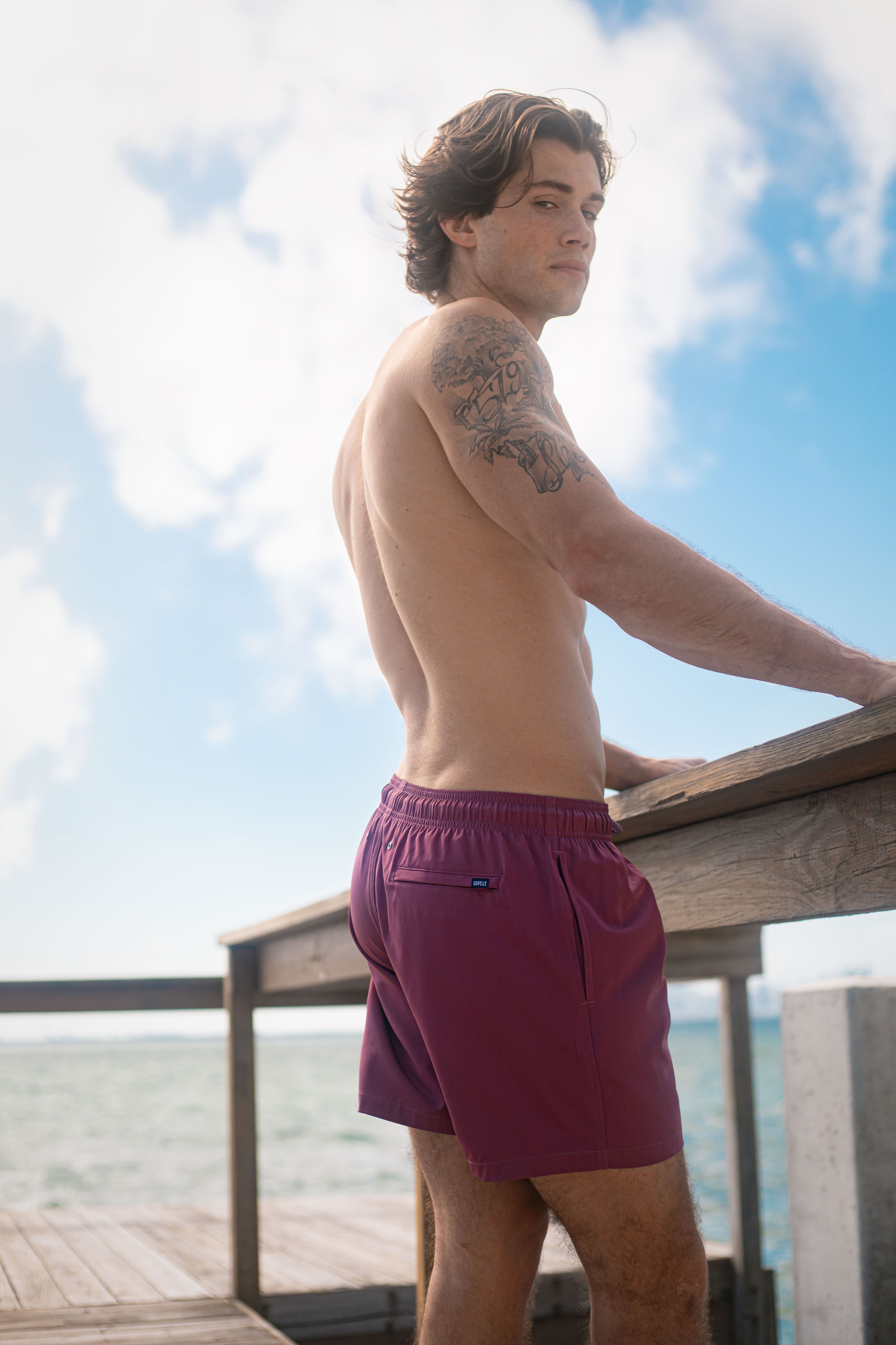 Man wearing purple swim shorts standing on a wooden deck with a blue sky and ocean background