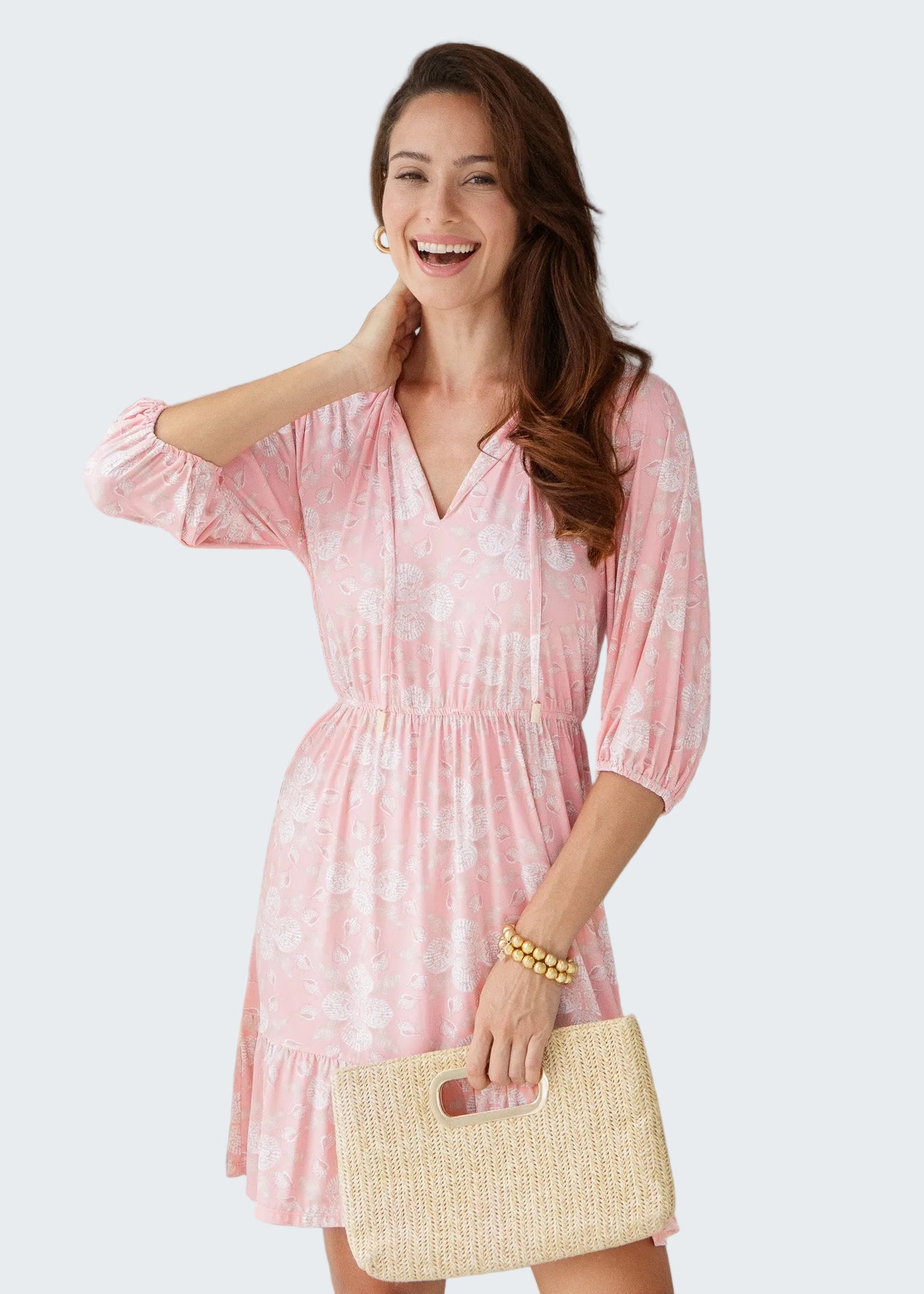 Woman wearing a pink dress with a white pattern, holding a straw bag indoors.