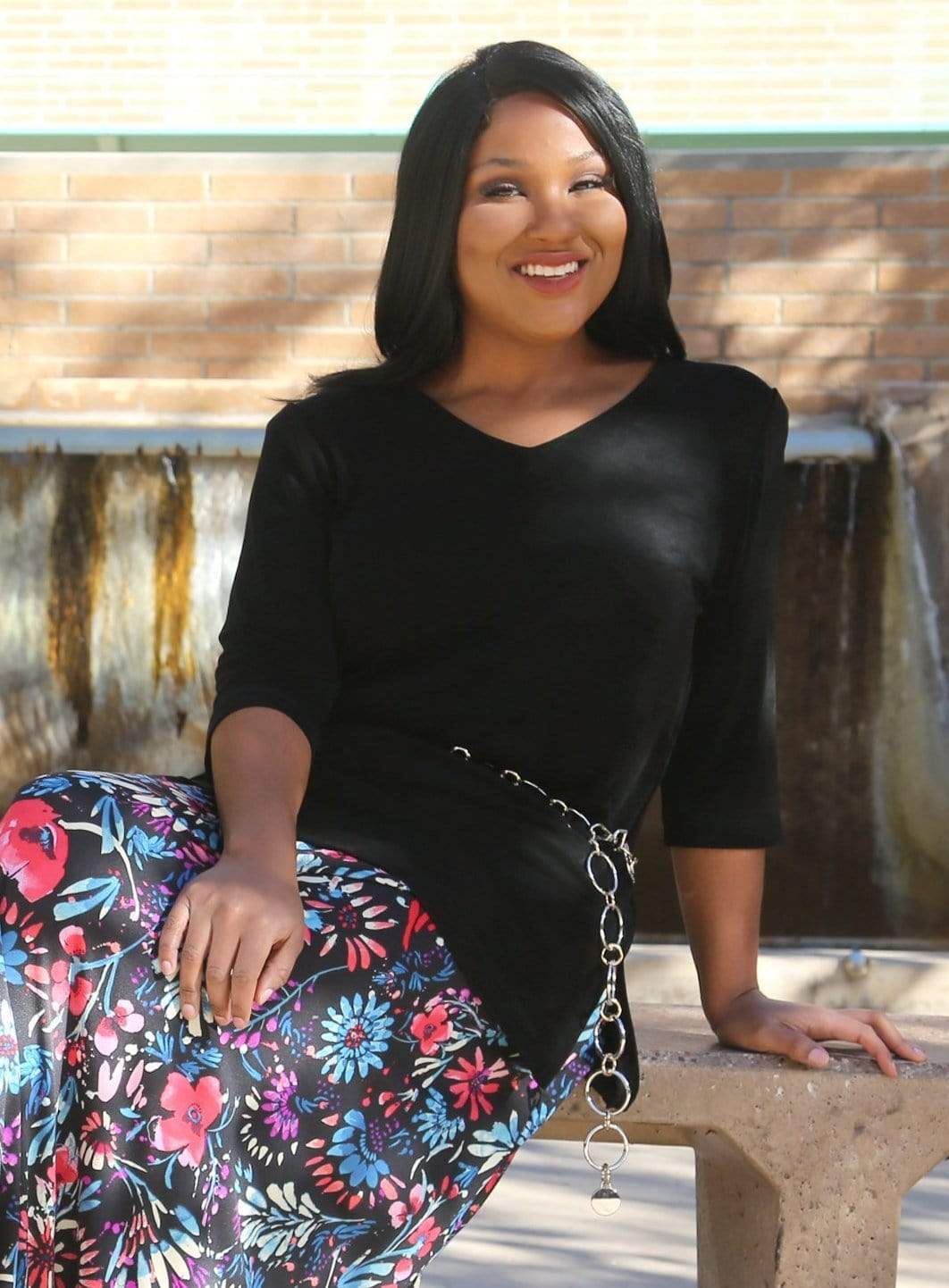 Woman wearing a black top and colorful floral skirt sitting on a bench.
