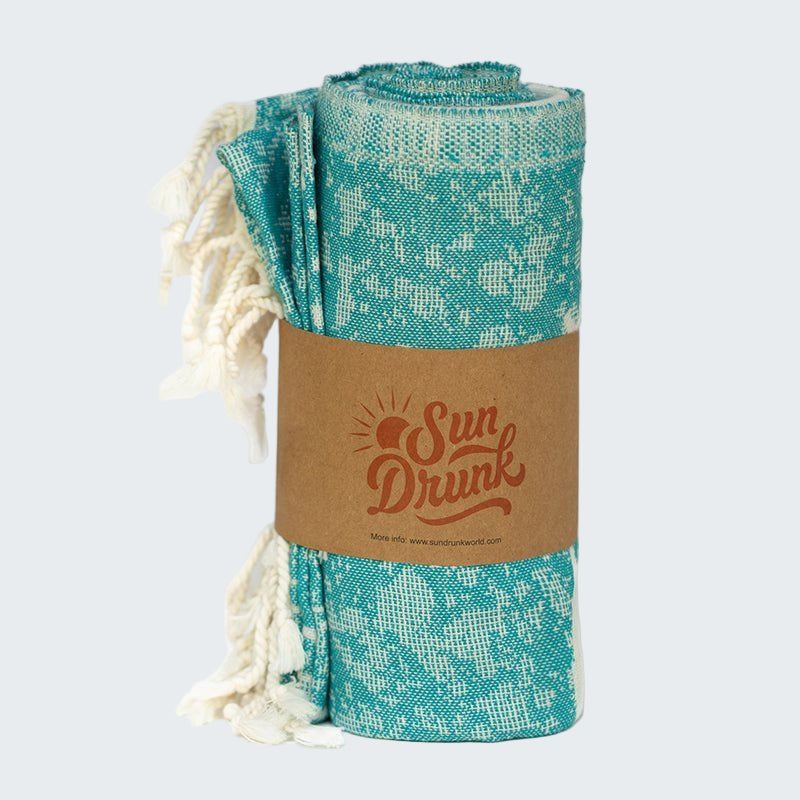 Teal patterned towel with white tassels on a cardboard label with 'Sun Drunk' branding.
