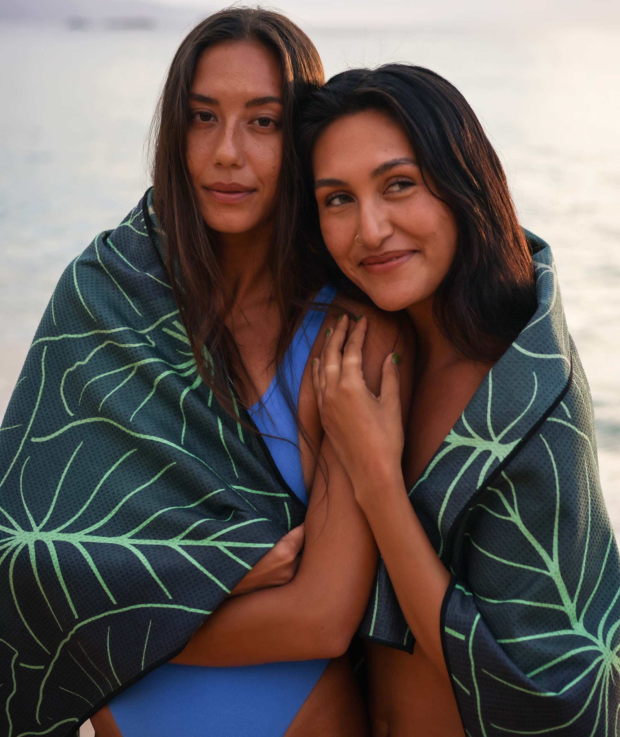 Two women wrapped in a green leaf-patterned towel by a body of water.