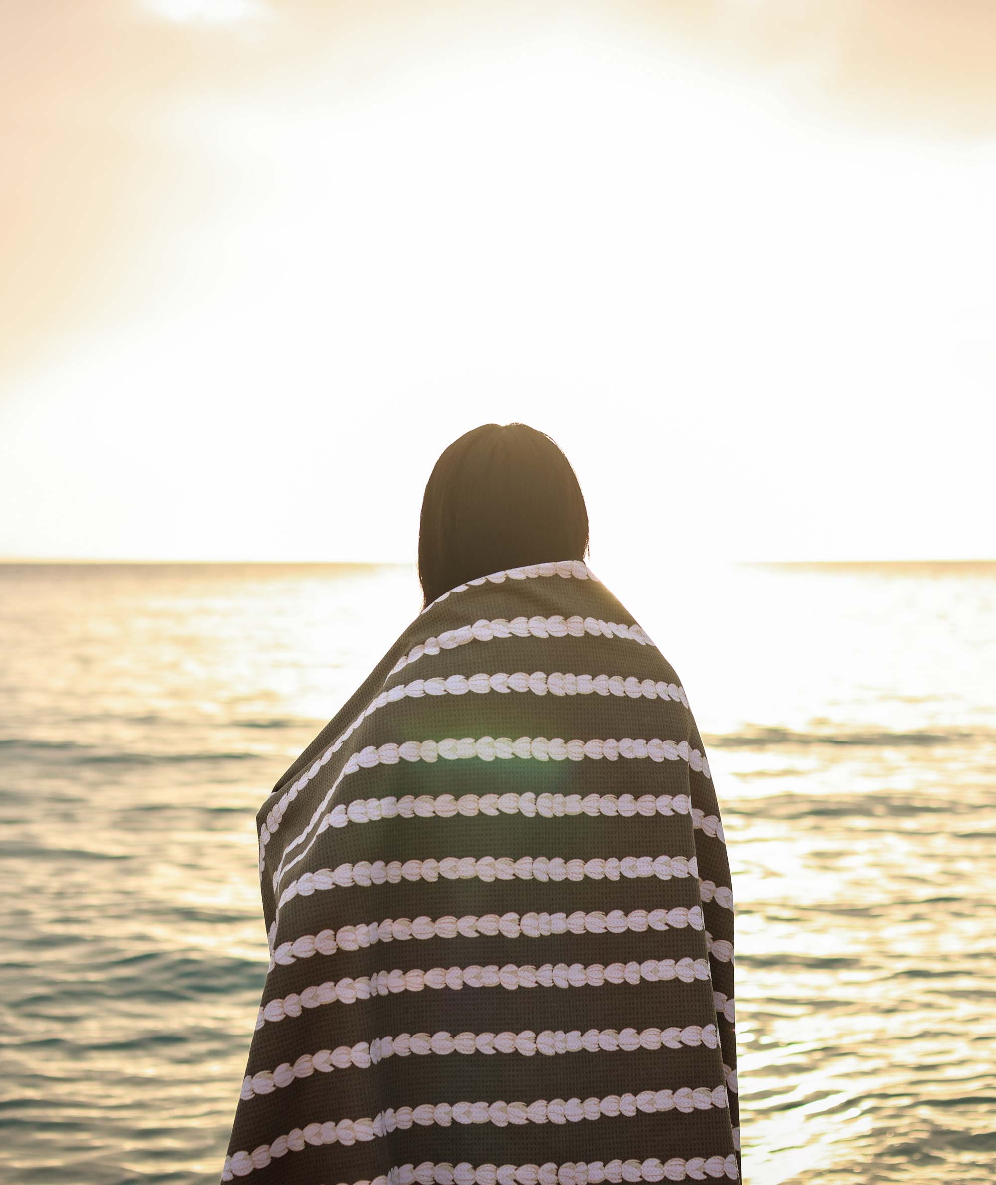 Person wrapped in a striped towel standing by the water at sunset