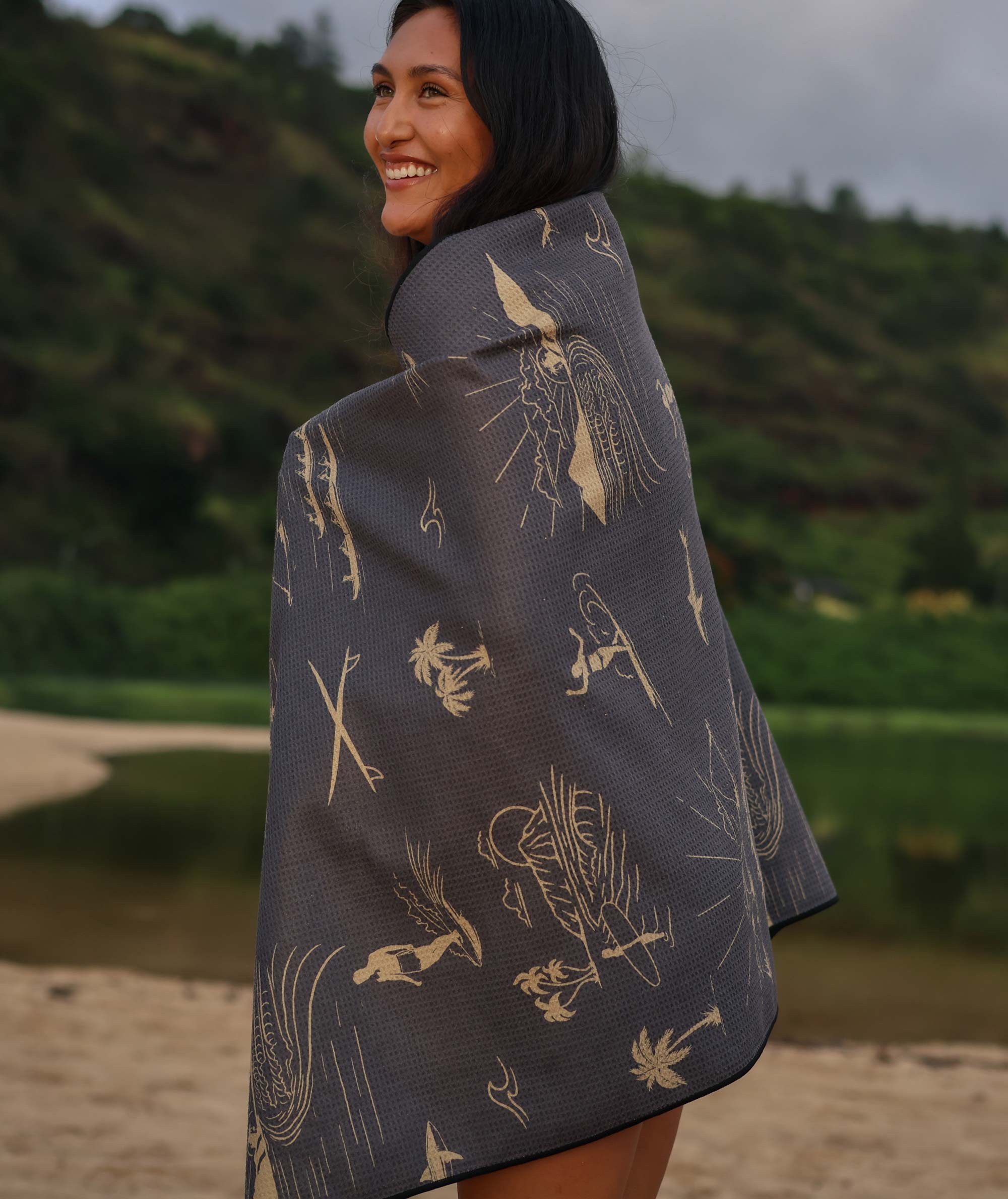Woman wearing a dark blue patterned poncho on a beach with greenery in the background