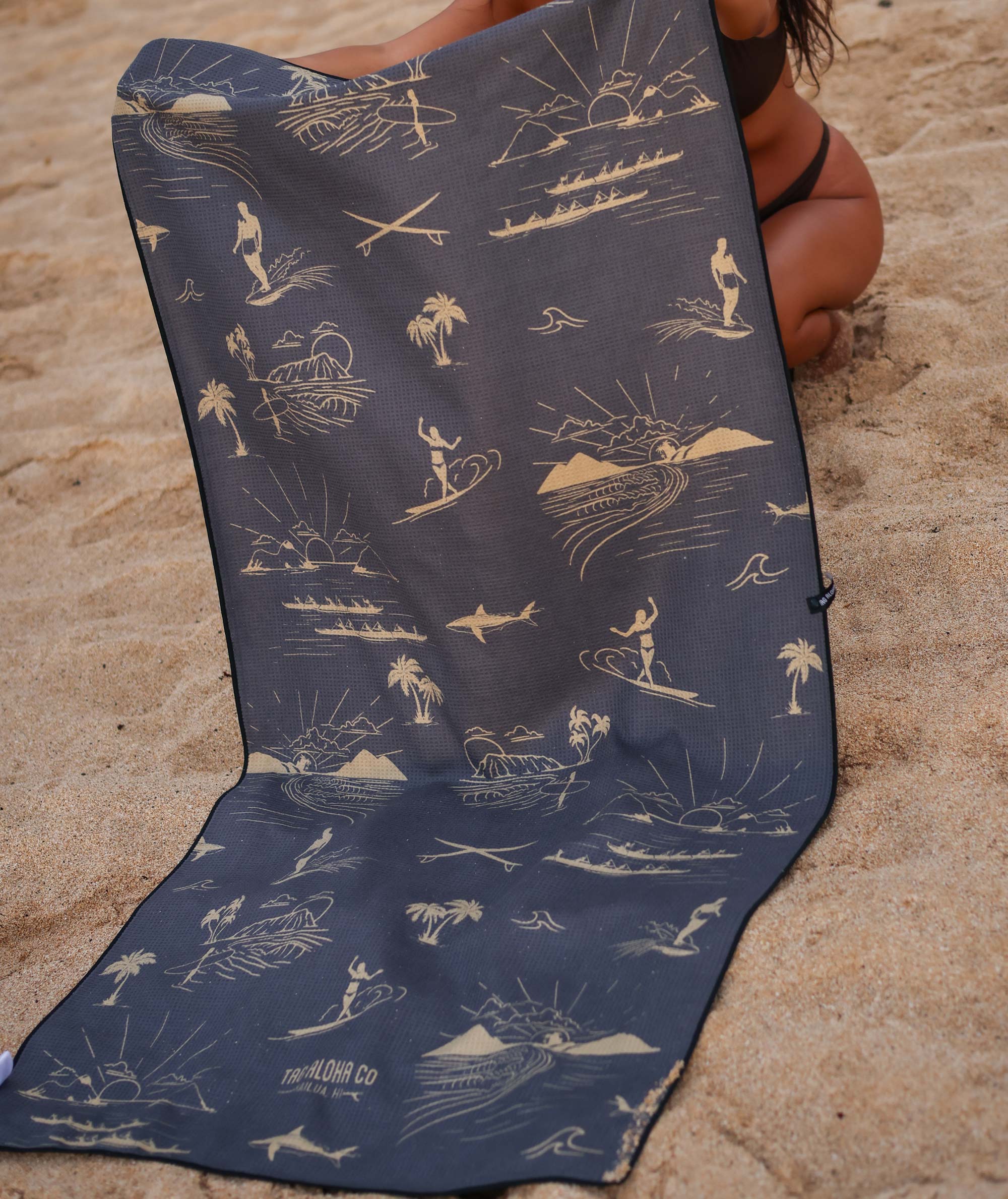 Navy blue beach towel with white beach-themed illustrations on a sandy background