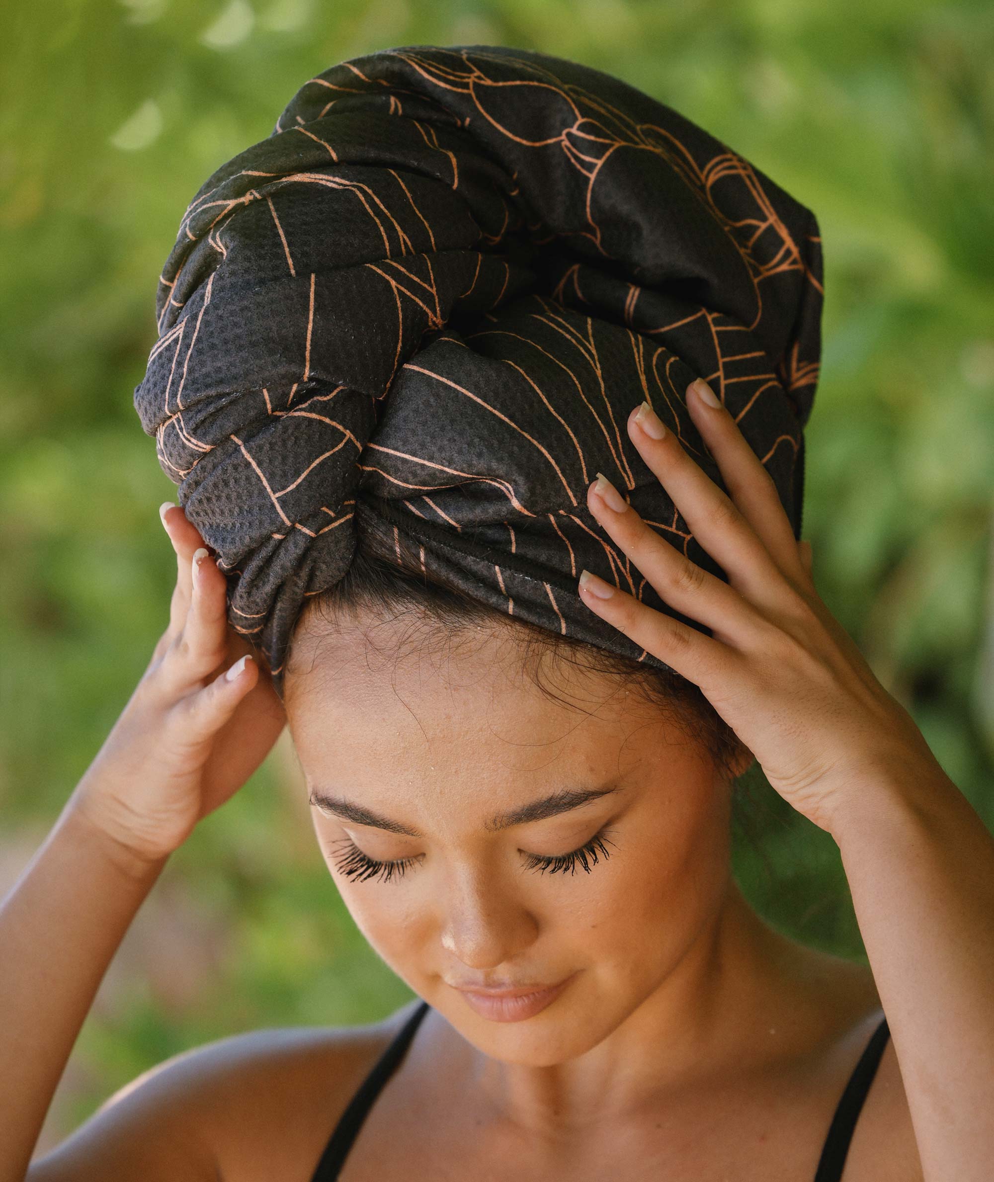 Woman with a black headscarf with gold patterns, standing outdoors with greenery in the background.