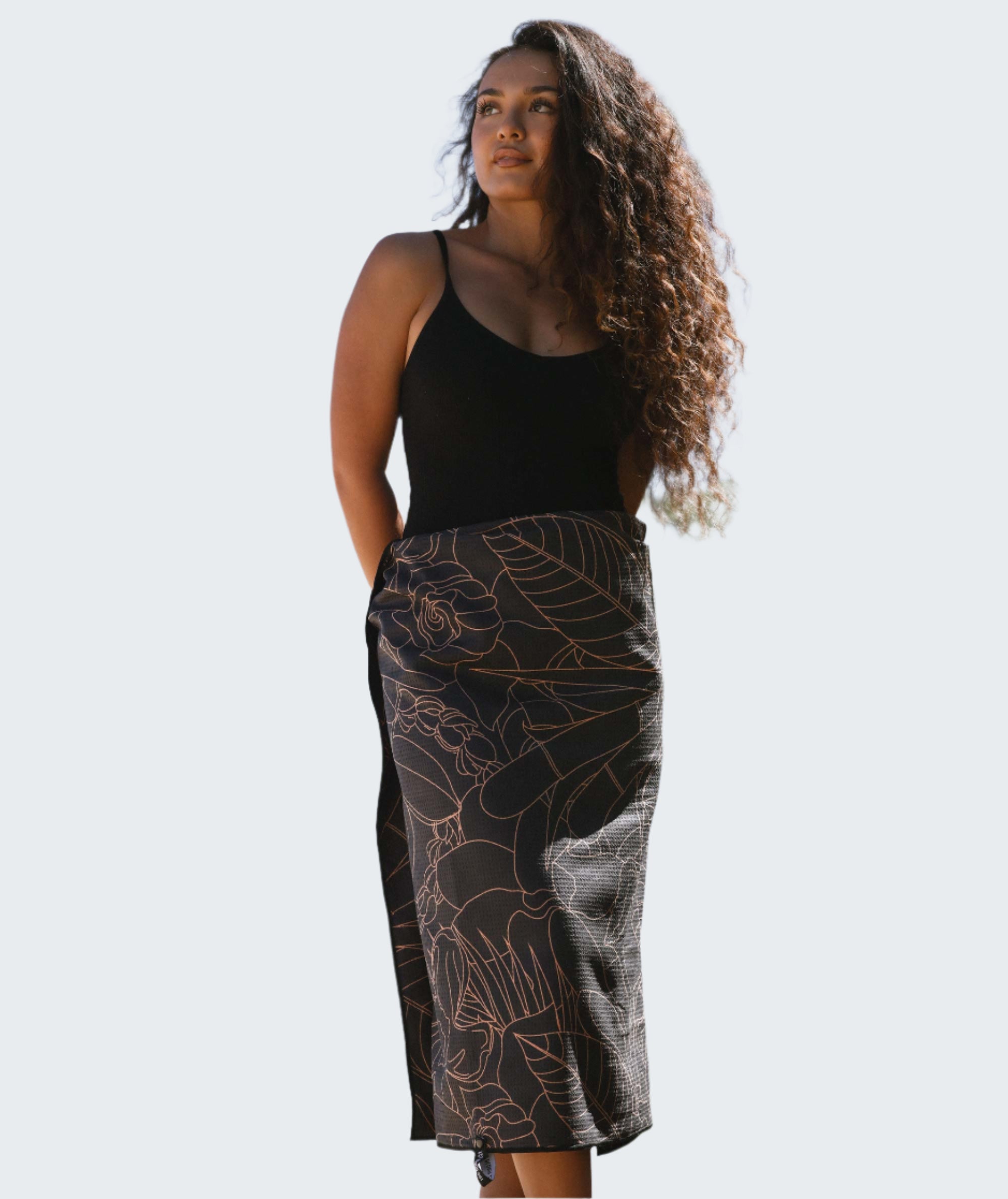 Woman wearing a black sleeveless top and patterned skirt outdoors with trees and blue sky in the background