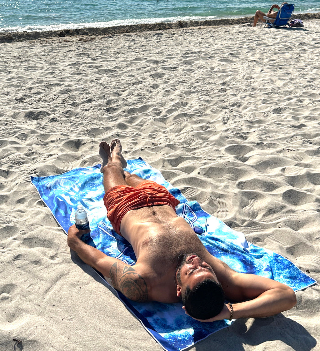 Man lying on a blue towel at the beach with ocean and other beachgoers in the background.