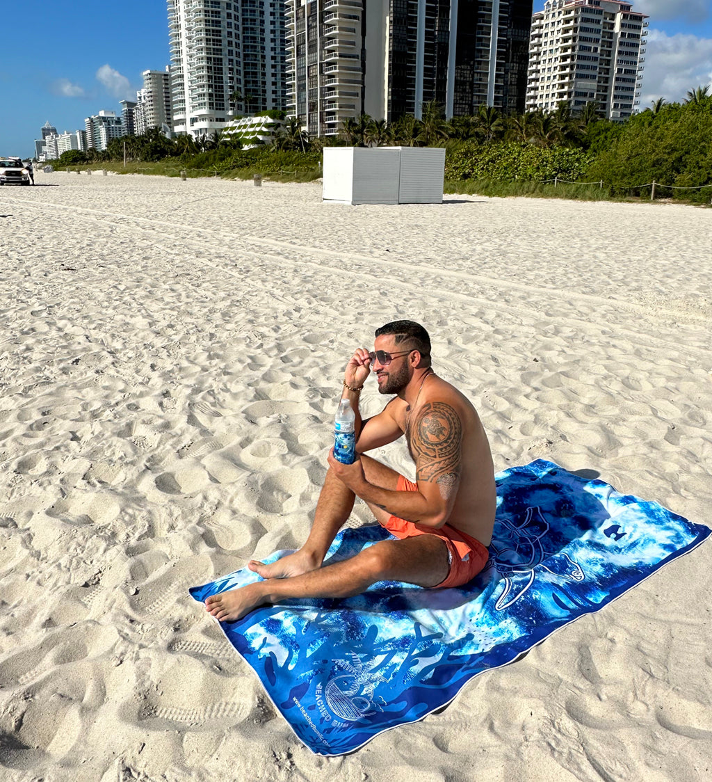 Man sitting on a blue towel on a sandy beach with buildings in the background