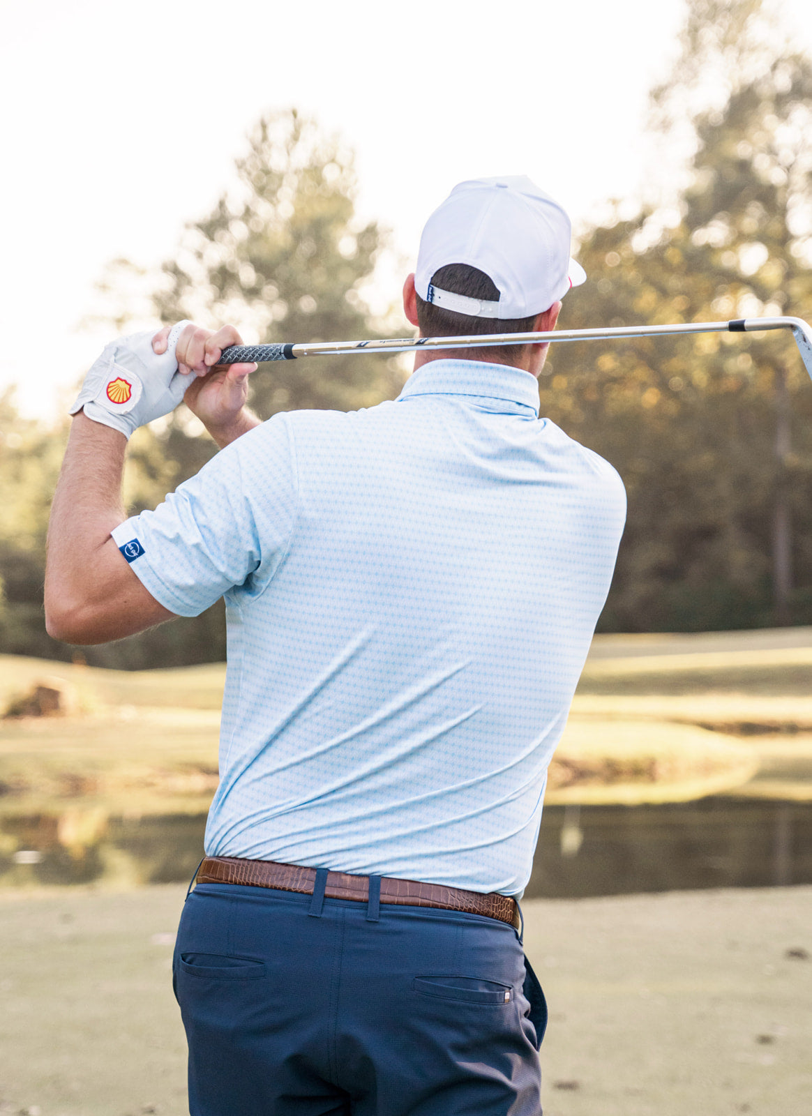 Golfer in a white cap and light blue shirt swinging a golf club on a golf course.