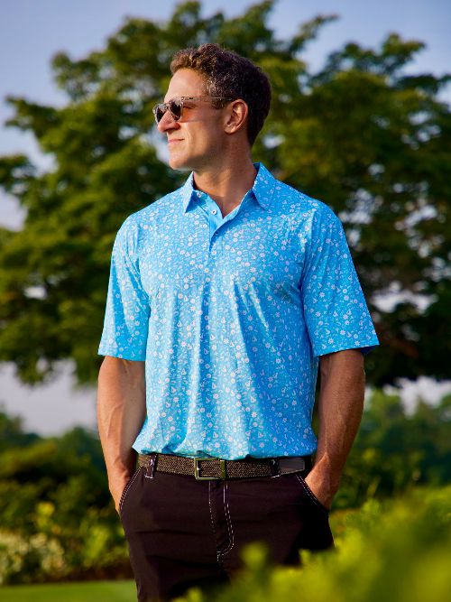 Man wearing a blue patterned shirt standing outdoors with greenery in the background