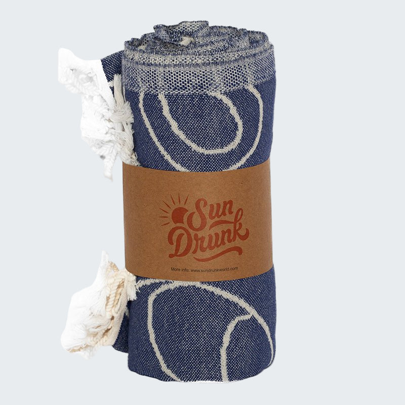 Rolled-up blue towel with white patterns and a brown strap labeled 'Sun Drunk' on a white background