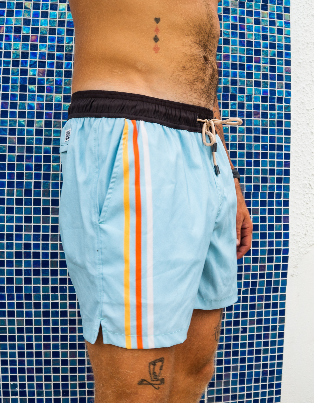Light blue swim shorts with colorful stripes worn by a person against a blue tiled wall.