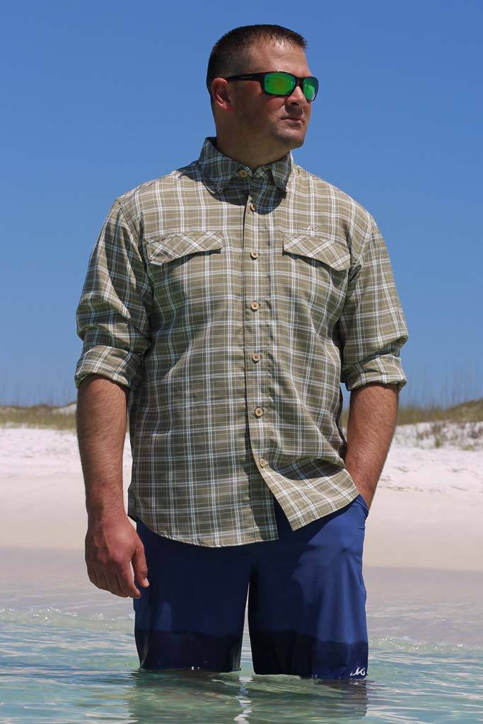 Man wearing a plaid shirt and sunglasses standing in shallow water on a beach.