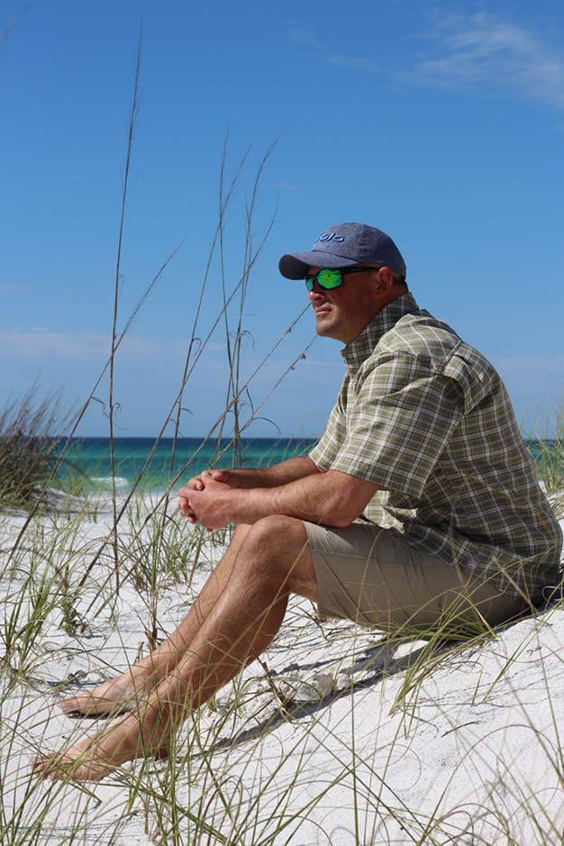 Man sitting on a sandy beach with grass, wearing sunglasses and a cap.
