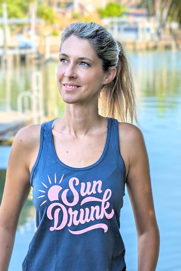 Woman wearing a blue tank top with 'Sun Drunk' text by a body of water.