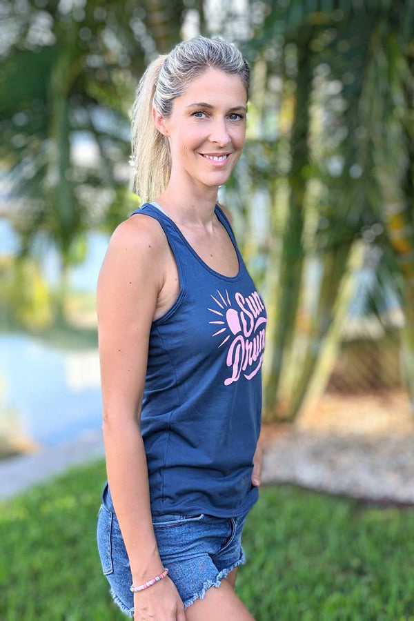 Woman wearing a navy tank top with a logo, standing outdoors with greenery in the background