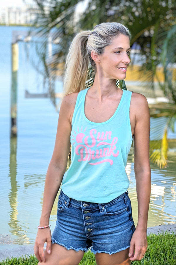 Woman wearing a light blue tank top with pink text and denim shorts by a water body.