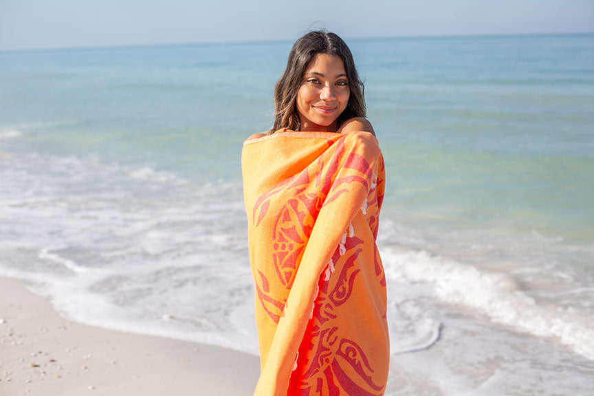 Woman in an orange and red patterned cover-up standing on a beach with ocean waves in the background.