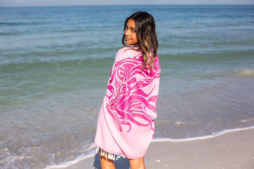 Woman standing on a beach with a pink patterned towel draped over her shoulders.