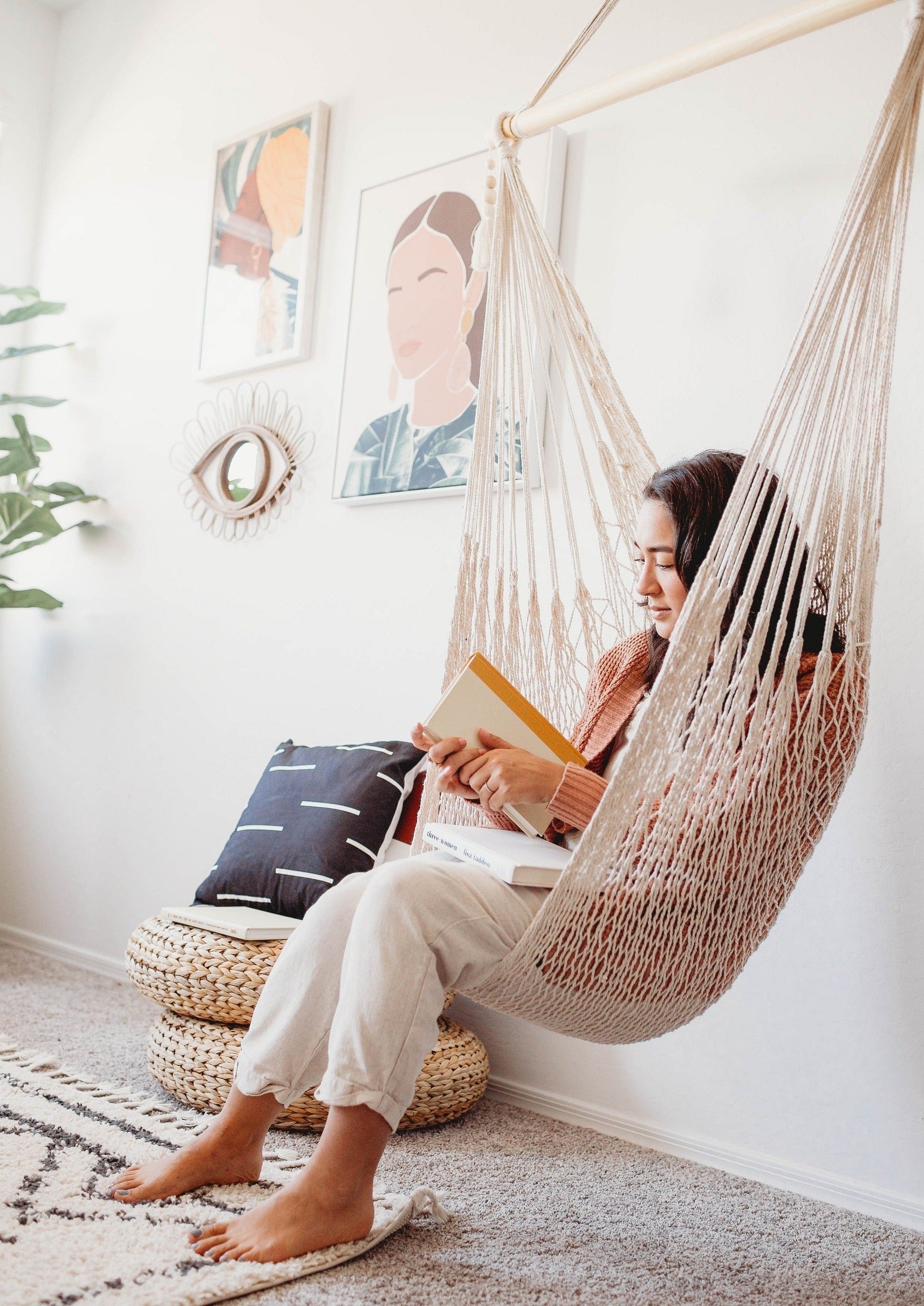 Person sitting in a hammock reading a book in a cozy room with decor elements.