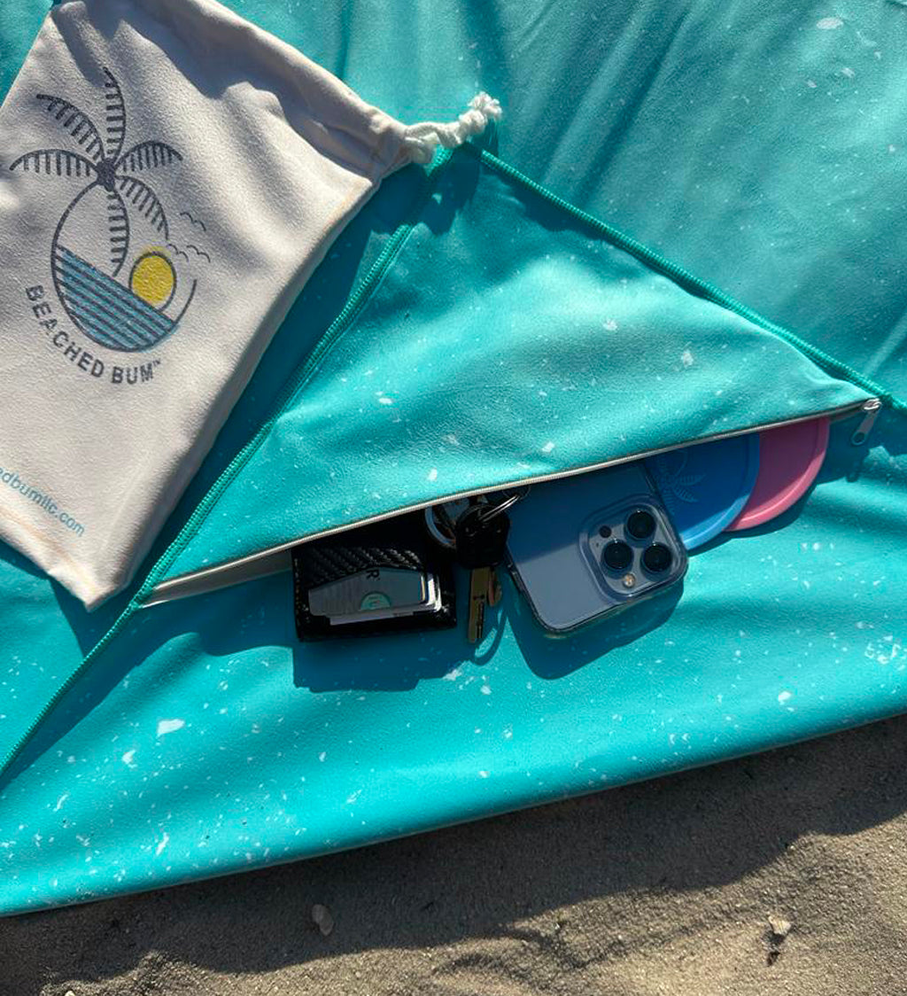 Teal towel with a phone and pouch on sand, featuring 'Beached Bum' logo.